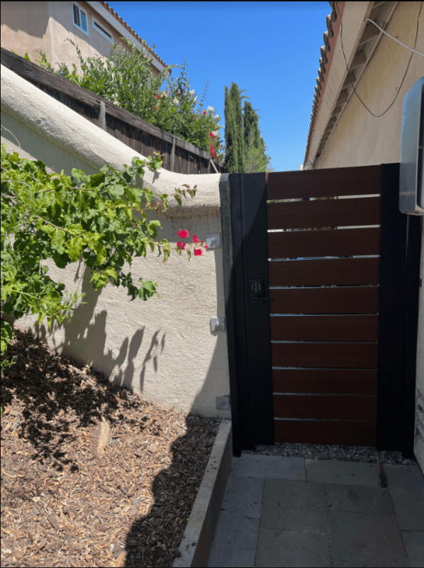 A newly installed fence with a wooden gate is shown. The gate features horizontal slats and is painted black, while the surrounding wall is light-colored stucco. To the left, there is a green bush with pink flowers, and the area is landscaped with mulch and stone paving. The sky is clear and blue, indicating a sunny day.