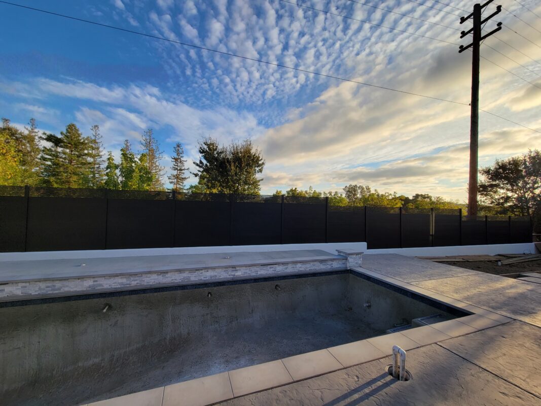 A modern black aluminum fence installed beside an empty pool under a cloudy sky.