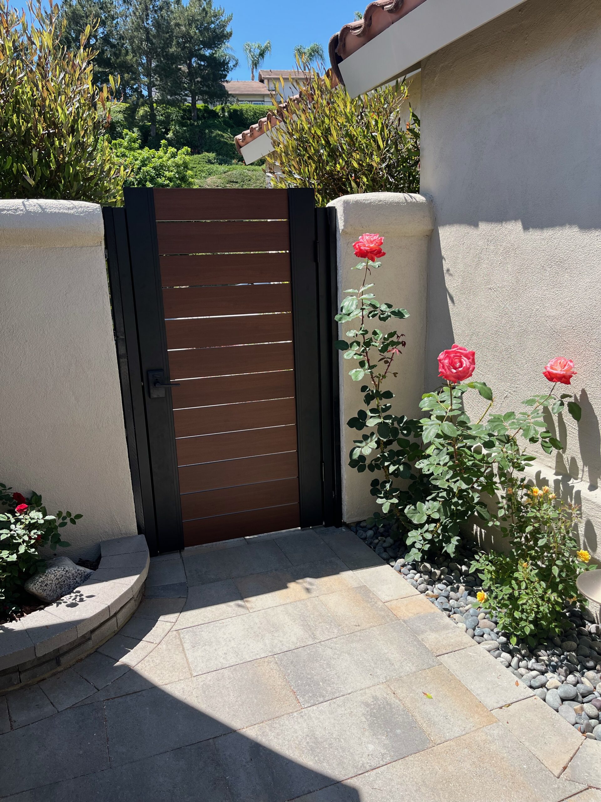 A newly installed wooden fence gate is set between two white concrete walls. The gate features a modern design with horizontal slats and is painted in a dark color. Flanking the gate are blooming pink roses and lush greenery, adding a touch of color to the entrance. The pathway leading up to the gate is paved with square gray stones, and there are small pebbles and plants along the edges. In the background, there are trees and houses visible under a clear blue sky.