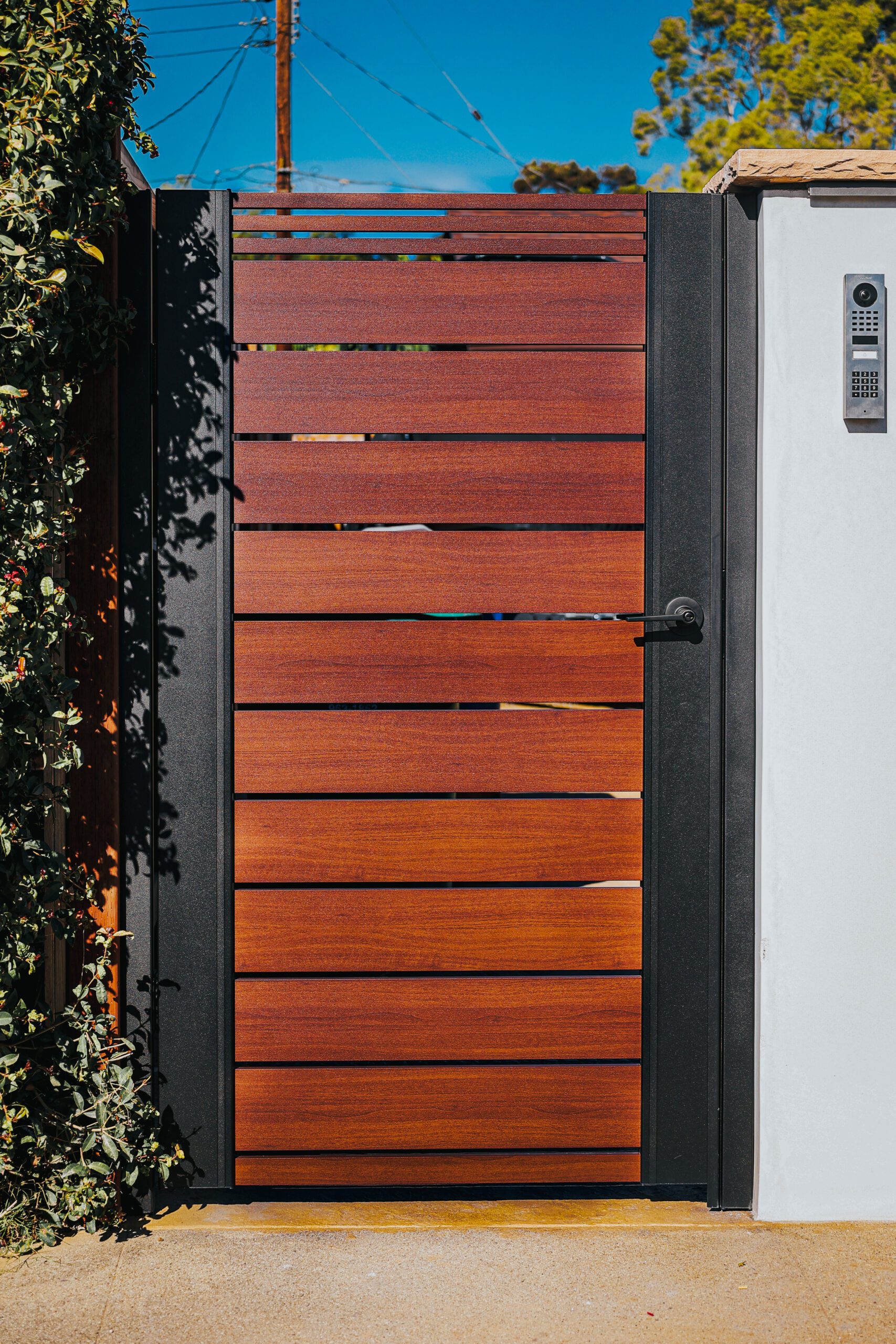Modern aluminum fence gate with wood-like finish, surrounded by greenery.
