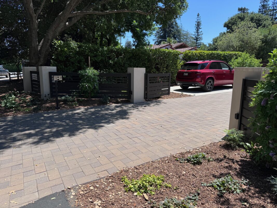 A stylish bronze aluminum gate and fence enhance a driveway with lush greenery.