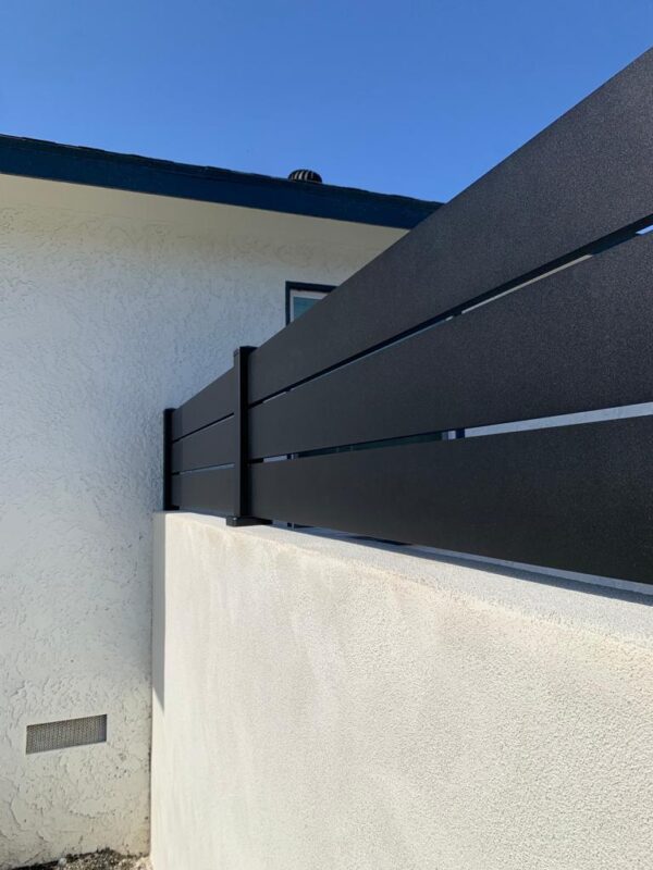 Modern black aluminum wall topper installed on a white stucco fence under a blue sky.
