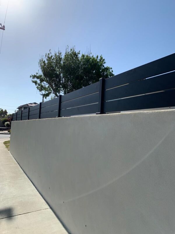 Newly installed black aluminum wall toppers atop a smooth gray wall under a clear sky.