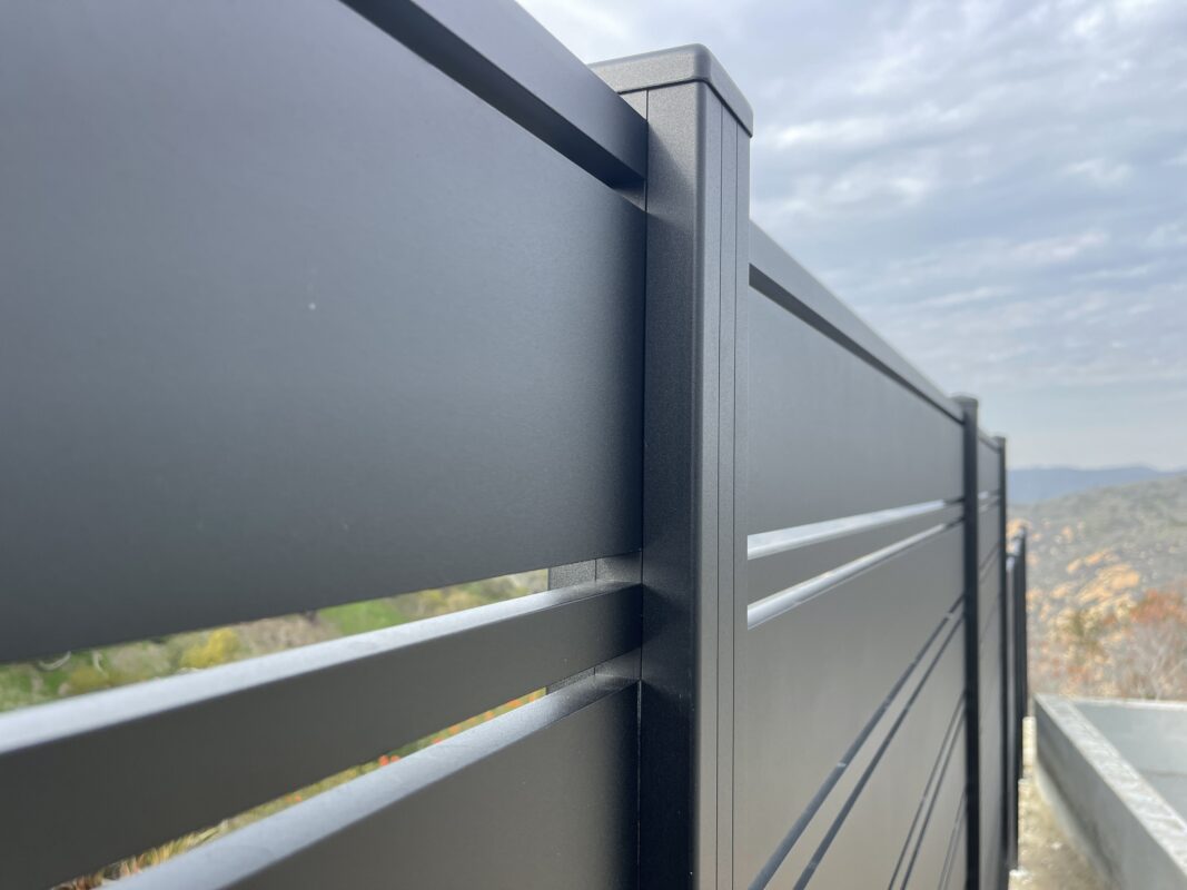 Close-up of a modern black aluminum fence on a hillside with cloudy skies.