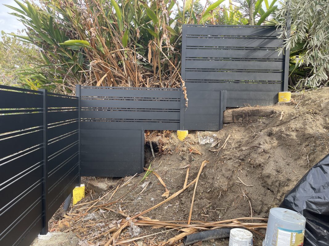 Close-up of a modern black aluminum fence installed on sloped terrain, surrounded by greenery.