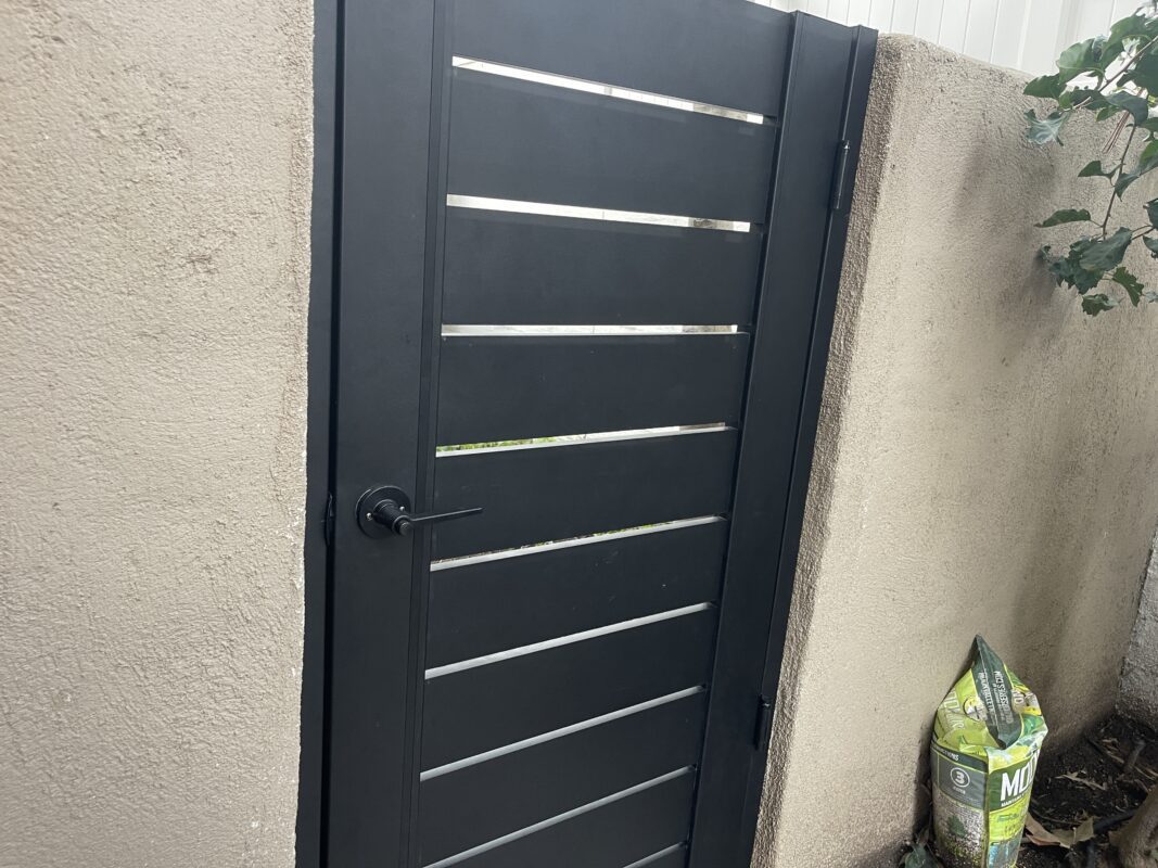 Modern black aluminum gate installed between two walls, partially obscured by foliage.