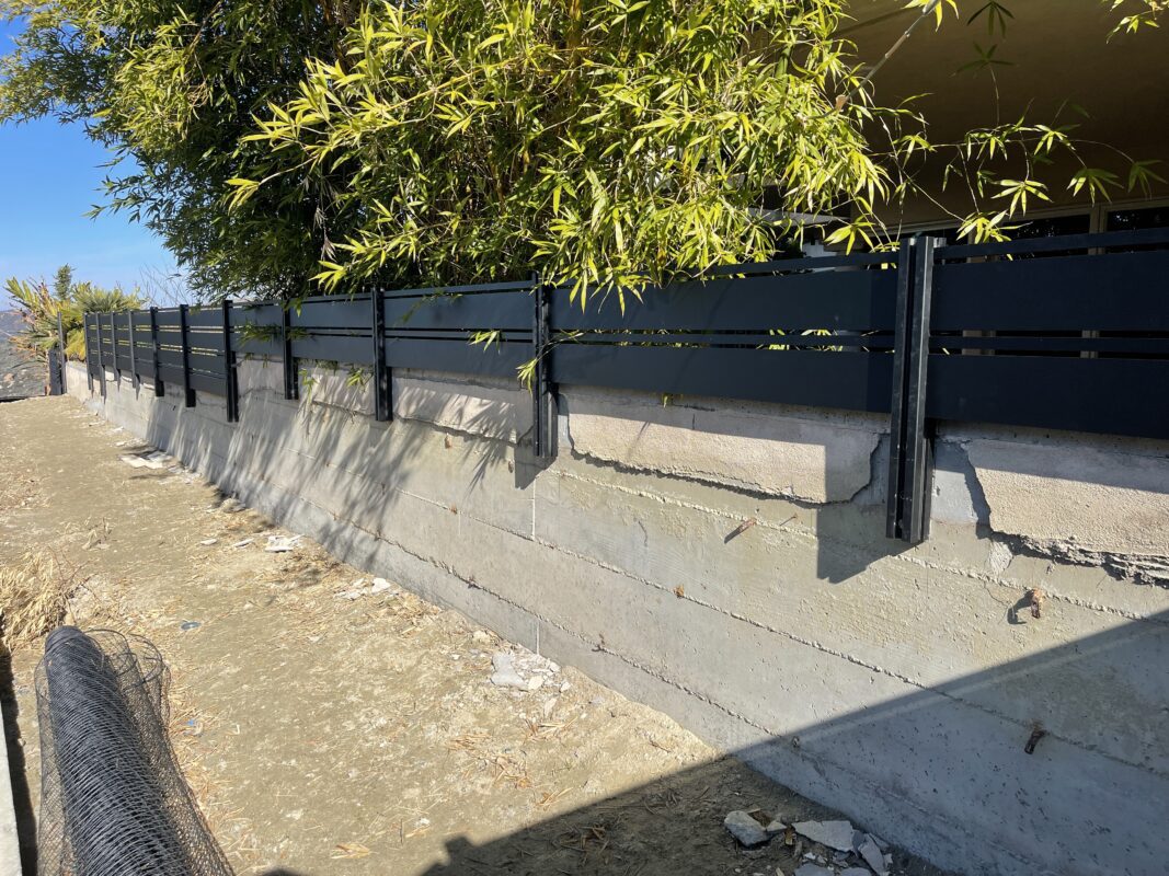 Modern black aluminum fence installed along a concrete wall in a sunny outdoor setting.