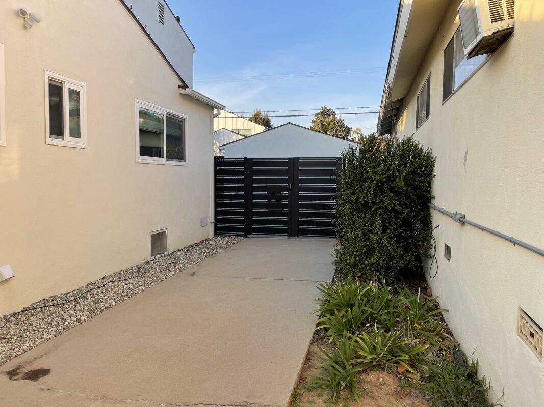 A black aluminum double swing gate installed between two buildings in a landscaped area.