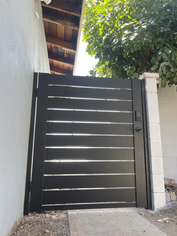 Black aluminum side gate installed next to a wall, surrounded by greenery and a pathway.