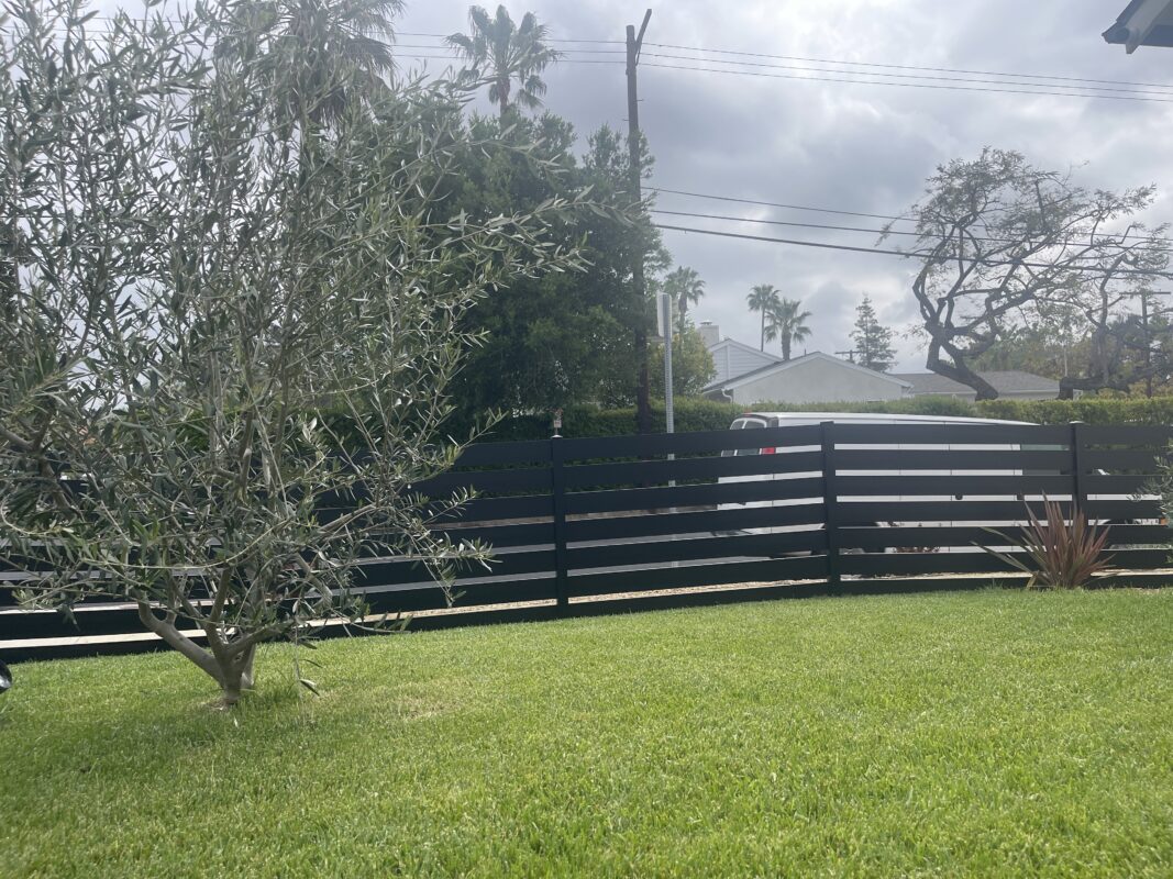 A modern black aluminum fence installed in a front yard with green grass and olive trees.