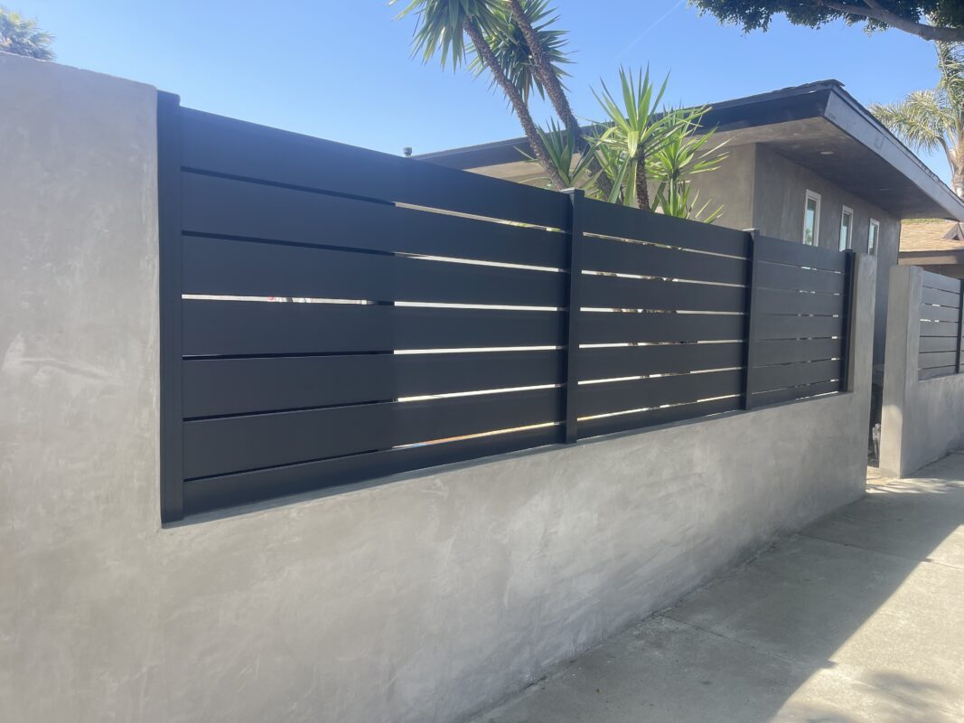 Modern black aluminum fence installed along a concrete wall in a sunny residential area.