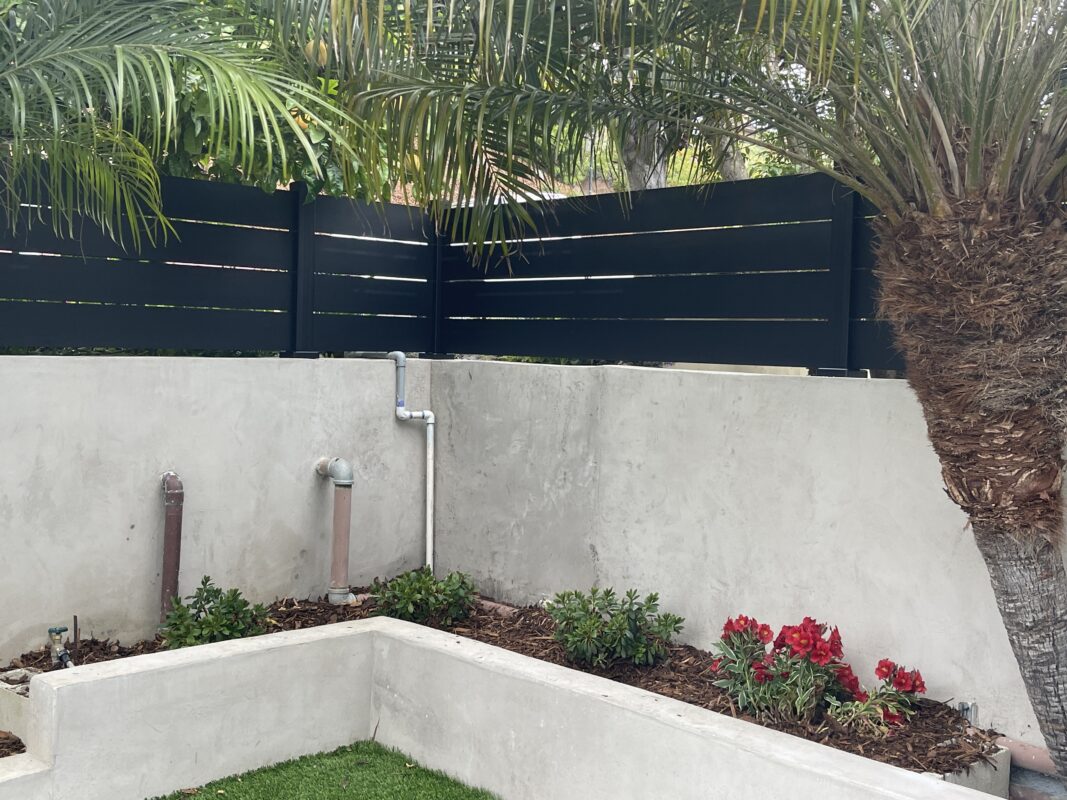 Modern black fence atop a concrete wall, surrounded by greenery and flowers.