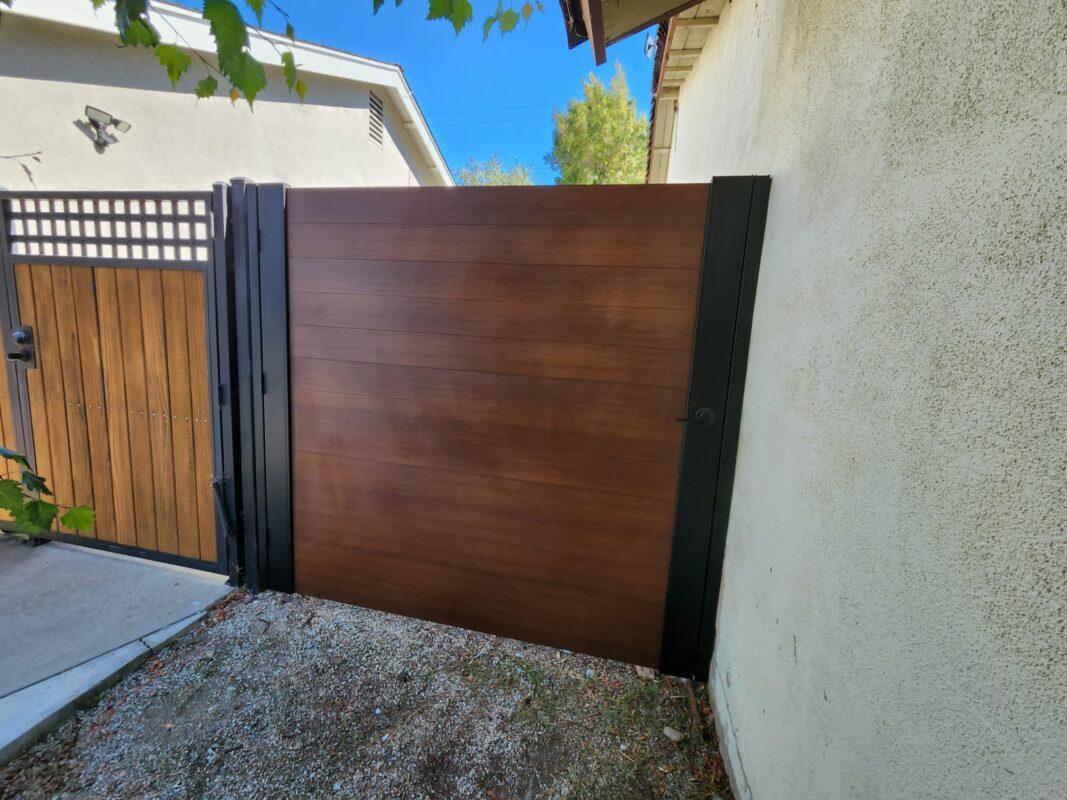 A sleek full privacy gate resembling wood, framed in black aluminum, installed beside a wall.
