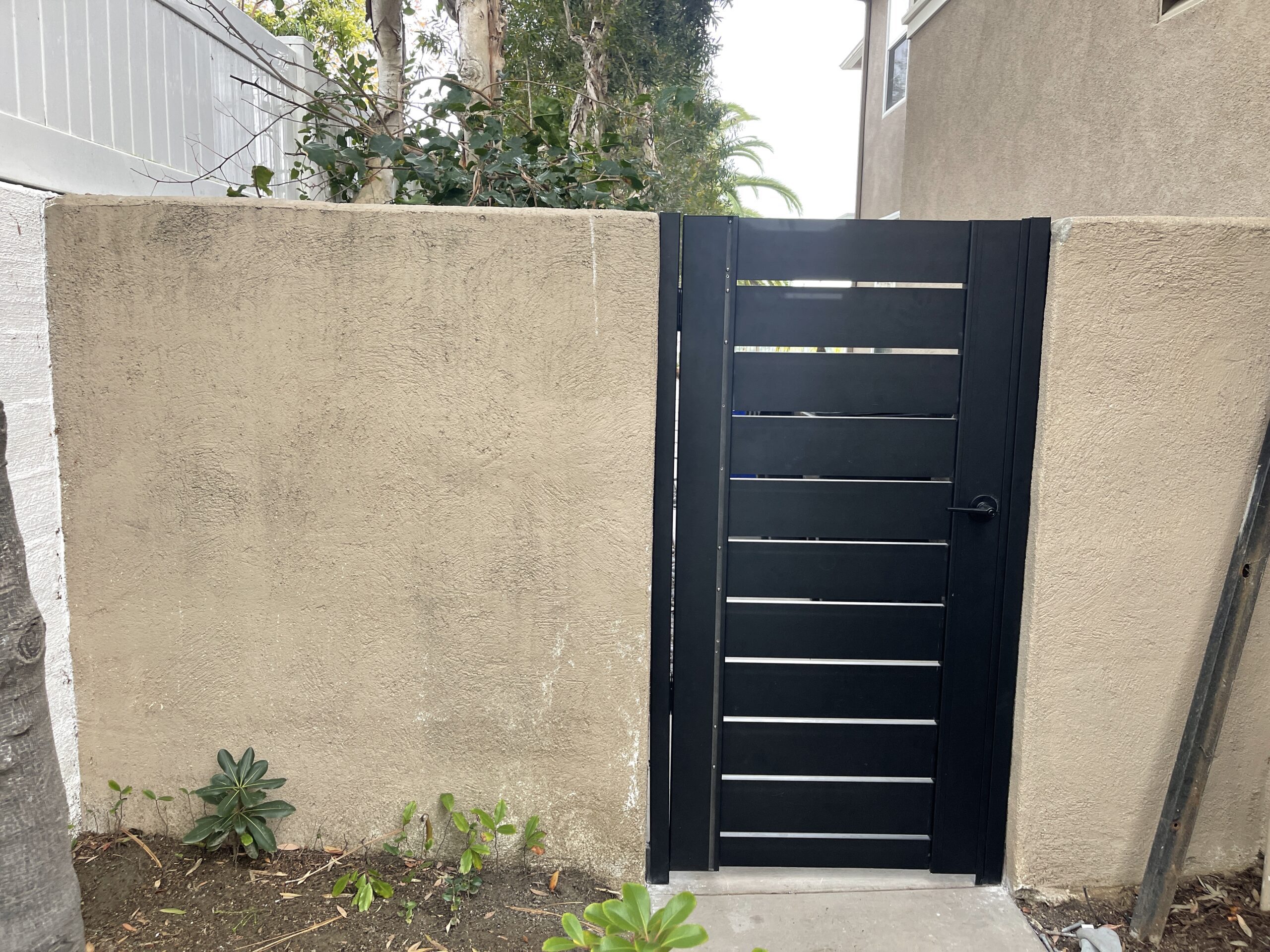 Black aluminum gate installed in a tan stucco wall with greenery around.