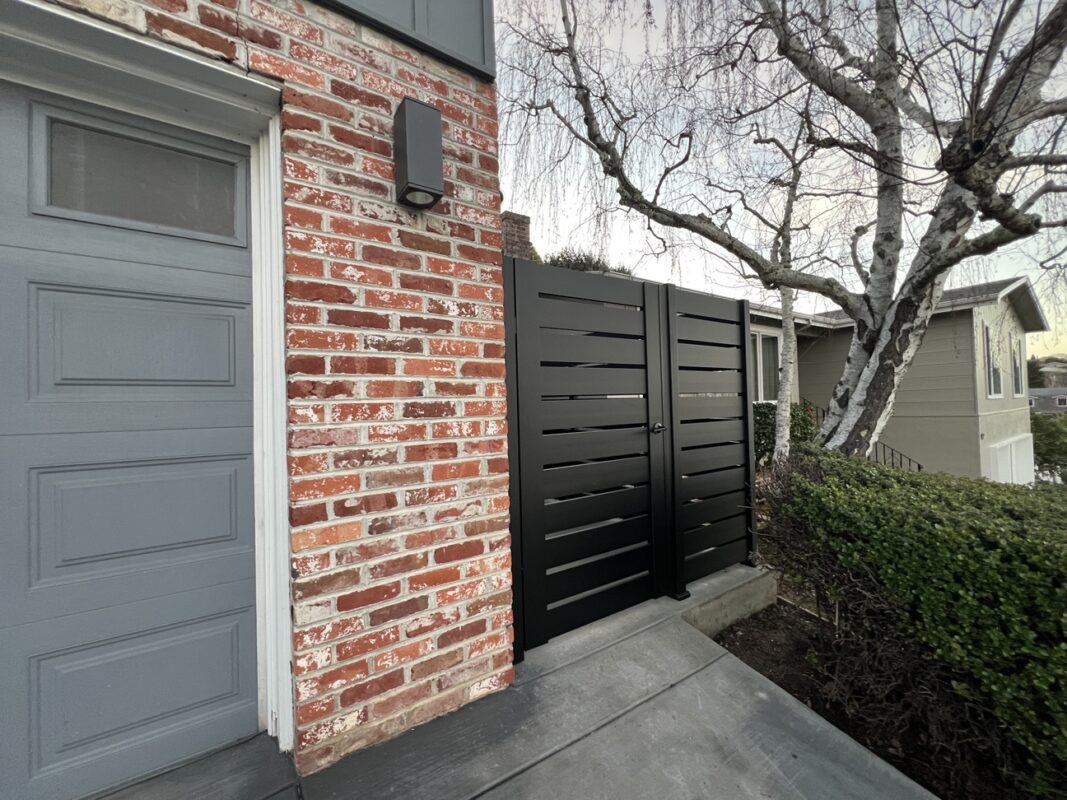 A sleek black pedestrian gate installed next to a brick wall and grey door.