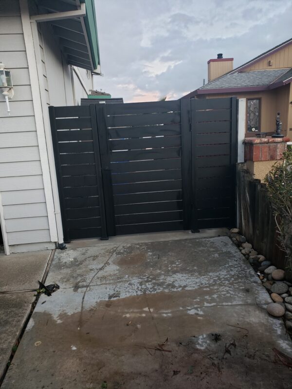 A modern black side yard gate installed next to a house, with visible concrete paving.