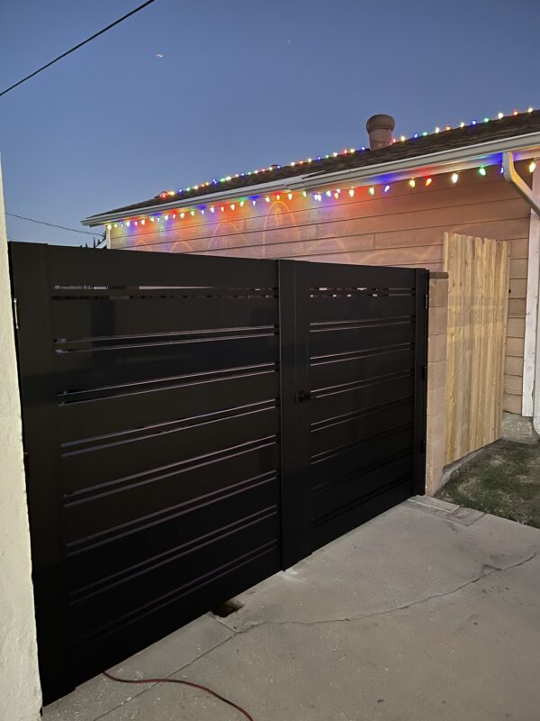 Black aluminum double swing gates installed next to a lit house in the evening.